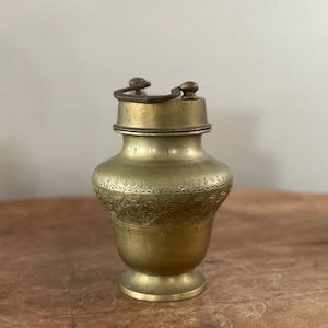 May include: A brass vessel with a wide, rounded body and a narrow neck, featuring an engraved band of repeating floral and geometric patterns. The container includes a fitted lid with a dark metal handle attached to two small, rounded knobs.