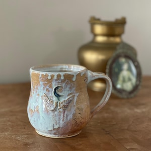 May include: A handmade ceramic mug with a moth and crescent moon design. The mug features a brown and white glaze with a blue accent. The handle is curved, and the mug has a rustic, artisanal aesthetic.