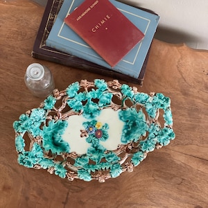 May include: An oval, ceramic serving dish with a leaf and vine design in shades of green and brown. The center features a cream-colored panel with a floral arrangement. A small glass bottle and books are also visible.