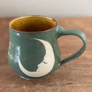 May include: Handmade ceramic mug in a teal green glaze with a cream-colored crescent moon design. The interior is a warm yellow-brown. The mug has a curved handle and is suitable for hot or cold beverages.