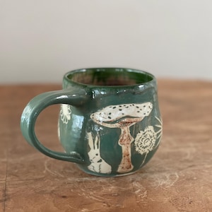 May include: A handcrafted ceramic mug with a dark green glaze. The mug features white illustrations of a mushroom, rabbit, and flowers. The handle is curved and the mug sits on a wooden surface.
