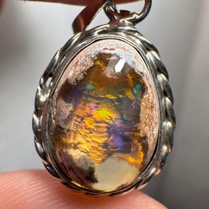 May include: A silver pendant with a large, oval-shaped opal gemstone. The opal has a white base with flashes of yellow, orange, green, and blue.