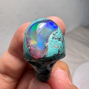 Könnte beinhalten: Ein einzigartiger Edelstein mit einem lebendigen, schillernden Opalzentrum, das ein Farbspektrum von Blau, Grün und Rot aufweist. Der Opal ist in einer türkisfarbenen Matrix eingebettet, was einen auffälligen Kontrast erzeugt. Der Stein hat eine raue, organische Form.