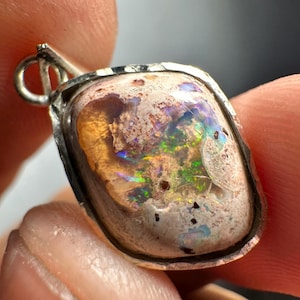 May include: A silver pendant with a light brown, white, and iridescent opal stone. The stone has a rough, textured surface and is set in a silver bezel.