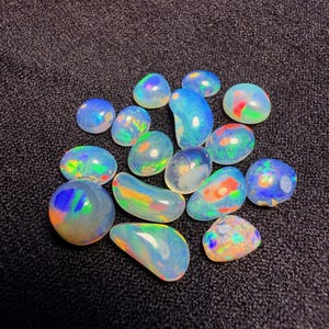 May include: A collection of polished opals in various shapes and sizes. The opals display a range of colors, including blues, greens, reds, and yellows, with iridescent flashes. The gemstones are set against a dark, textured background.