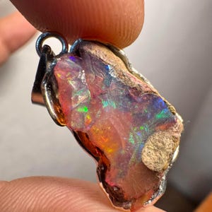 Op de afbeelding: Een zilveren hanger met een ruwe, iriserende opaal met een ruwe, natuurlijke textuur. De opaal vertoont een scala aan kleuren, waaronder roze, oranje, groen en blauw.
