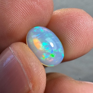 Puede incluir: Una piedra preciosa de ópalo ovalada con una base blanca lechosa. El ópalo muestra destellos iridiscentes de tonos verdes, azules y naranjas en su superficie. La gema pulida se sostiene entre dos dedos, resaltando su acabado brillante.