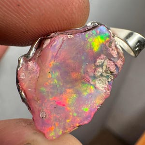 May include: A pink opal pendant with a silver setting. The opal has a rough, natural shape and displays a variety of colors, including pink, green, and orange.
