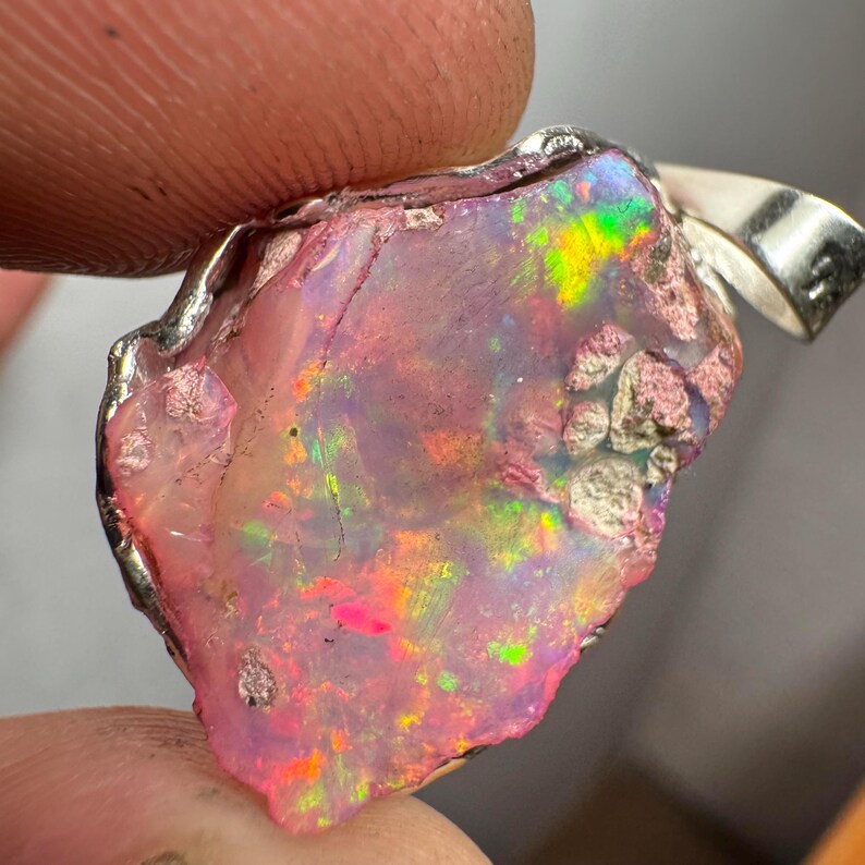 May include: A pink opal pendant with a silver setting. The opal has a rough, natural shape and displays a variety of colors, including pink, green, and orange.