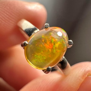 Puede incluir: Un anillo de plata con una piedra preciosa de ópalo de forma ovalada. El ópalo presenta un brillo iridiscente amarillo, verde y naranja.