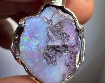 May include: A silver pendant featuring a large, irregular-shaped opal with a purple, blue, and green iridescent sheen. The opal is set in a silver frame with a textured finish.