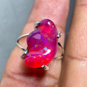 May include: A silver ring featuring a large, pink opal gemstone. The opal has a distinctive, freeform shape and showcases a range of colours, including pink, purple, and blue.