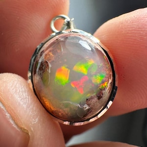 May include: A silver pendant with a round, iridescent opal stone. The opal has flashes of green, red, and orange.