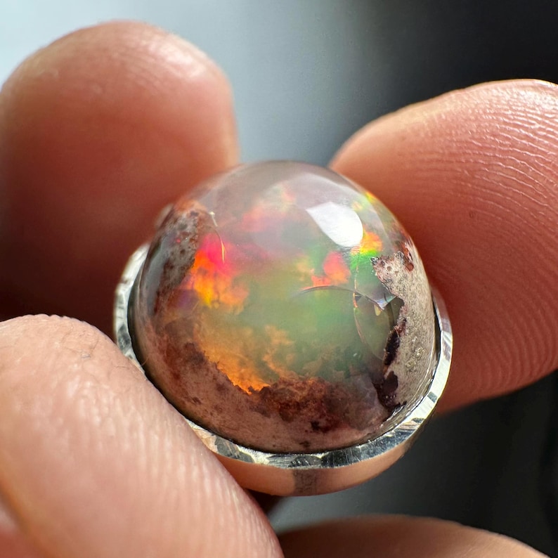 May include: A silver ring with a large, oval-shaped opal gemstone. The opal has a milky white base with flashes of red, orange, green, and blue.