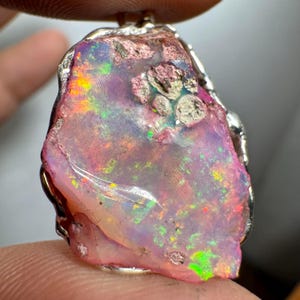 May include: A pink opal pendant with a silver setting. The opal has a variety of colors, including pink, orange, green, and blue. The opal is set in a rough, natural shape.
