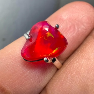 May include: A silver ring featuring a large, red, irregularly shaped opal gemstone. The opal displays a vibrant, fiery red hue with hints of yellow and pink.