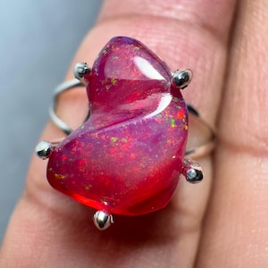 May include: A silver ring featuring a large, freeform, red opal gemstone. The opal displays a swirling pattern and flashes of green and purple.