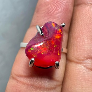 May include: A silver ring featuring a large, freeform, pink opal gemstone. The opal displays a vibrant, iridescent sheen with flashes of orange, red, and green.