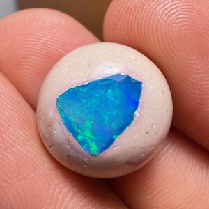 Puede incluir: Una piedra redonda y clara con una incrustación de ópalo azul y verde vibrante. El ópalo tiene una forma irregular llamativa y está engastado en la superficie lisa y pálida de la piedra. Los colores del ópalo cambian con la luz.