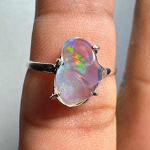 Puede incluir: Un anillo de plata con una gran piedra preciosa de ópalo iridiscente. El ópalo muestra una gama de colores, incluyendo rosa, verde y azul, con una forma orgánica única. La banda del anillo es de plata pulida.