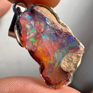 Op de afbeelding: Een ruwe, natuurlijke opaal hanger met een zilveren zetting. De opaal is een levendige roze, oranje en groene kleur met iriserende flitsen. De opaal is gezet in een zilveren bezel zetting.