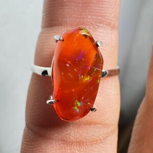 May include: A silver ring with a striking orange opal. The opal displays iridescent flashes of green, purple, and yellow. The gemstone is secured by four silver prongs. The ring's band is a simple, polished silver design.