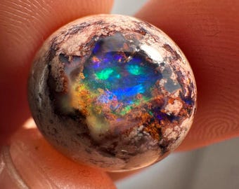 May include: A round, iridescent opal gemstone with a brown matrix. The opal displays a rainbow of colors, including green, blue, and purple.