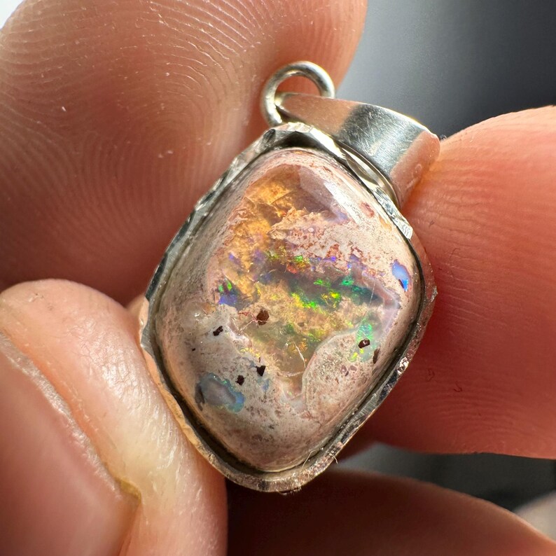 May include: A silver pendant with a light brown, white, and iridescent opal stone. The stone has a rough, textured surface and is set in a silver bezel.