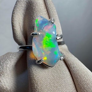 May include: A silver ring with a large, freeform opal gemstone. The opal exhibits a striking display of colour, including green, blue, and yellow hues. The ring has a plain silver band, and the opal is secured by four silver prongs.