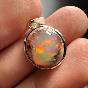 May include: A silver pendant with a round, iridescent opal gemstone. The opal has a mix of orange, red, green, and blue colors.