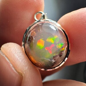 May include: A silver pendant with a round, iridescent opal gemstone. The opal has flashes of green, red, and orange.