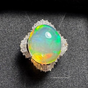 Puede incluir: Un anillo con una gran ópalo iridiscente. El ópalo muestra un vibrante juego de colores, incluyendo tonos verdes, amarillos y azules. El anillo está engastado en una montura de plata u oro blanco, rodeado de piedras preciosas claras más pequeñas.