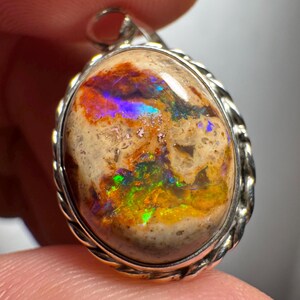 May include: Oval-shaped opal pendant with a silver-colored twisted rope border. The opal features a mix of colors, including orange, green, blue, and yellow, set against a beige background. The pendant has a silver loop at the top.