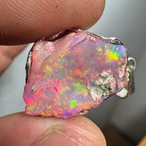 May include: A raw pink opal pendant with a silver setting. The opal has a rough, natural texture and displays a rainbow of colors, including pink, green, and blue.