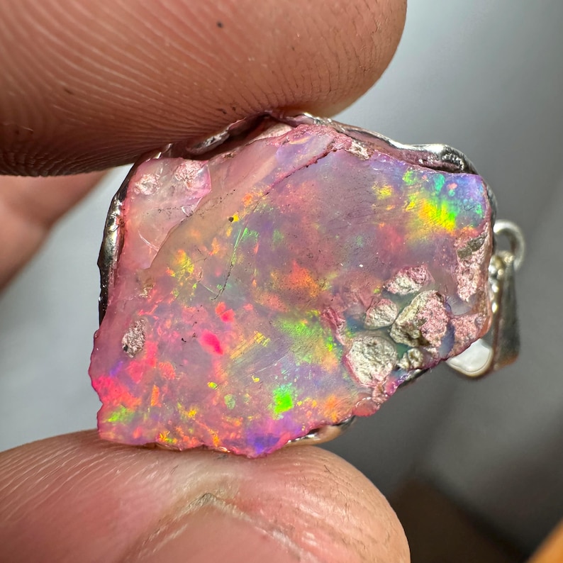 May include: A raw pink opal pendant with a silver setting. The opal has a rough, natural texture and displays a rainbow of colors, including pink, green, and blue.