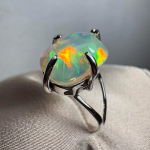 May include: A silver ring featuring a large, oval opal gemstone. The opal has a milky white base with flashes of green, orange, and yellow.