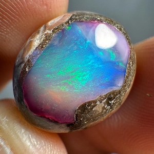 Puede incluir: Una piedra preciosa de ópalo pulida con una vibrante exhibición de colores. El ópalo presenta una mezcla de tonos azules, verdes y morados en su núcleo, enmarcado por una capa exterior rugosa y texturizada. La piedra se sostiene en una mano.