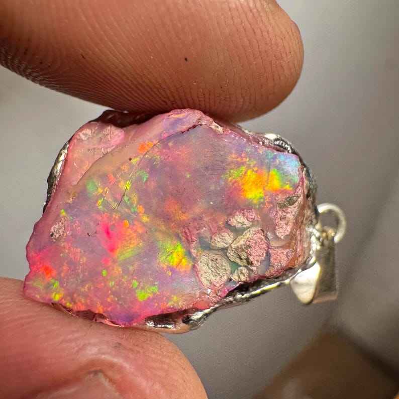 May include: A raw pink opal pendant with iridescent rainbow flashes, set in a silver bezel. The opal has a rough, natural texture and is a unique piece of jewelry.