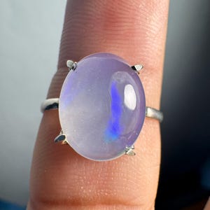 Puede incluir: Anillo de plata con una gran gema ovalada de color lavanda. La gema tiene una calidad translúcida con reflejos azules e iridiscentes. La banda del anillo es de plata pulida.