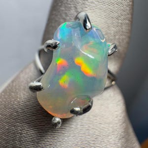 May include: A silver ring featuring a large, irregularly shaped opal gemstone. The opal is milky white with flashes of green, yellow, and orange.