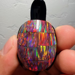 Puede incluir: Una figura de tortuga hecha a mano con cabeza y aletas negras. El caparazón de la tortuga está hecho de ópalo iridiscente multicolor, que muestra tonos de rojo, naranja, verde y morado. La figura se sostiene en la mano de una persona.