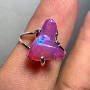May include: A silver ring featuring a large, freeform pink opal gemstone. The opal displays iridescent blue and green flashes.