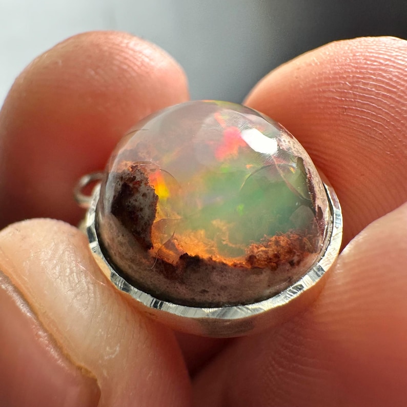 May include: A silver pendant with a round, clear opal stone. The opal has a brown, red, and green iridescent sheen.