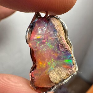 Op de afbeelding: Een ruwe, iriserende opaal hanger gezet in zilver. De opaal is een mix van roze, oranje, groen en blauw.