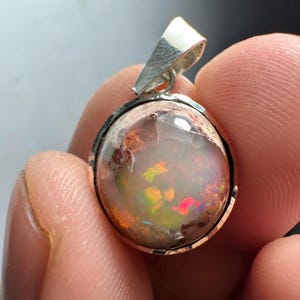 May include: A silver pendant with a round opal gemstone. The opal has a white base with flashes of orange, red, and green.