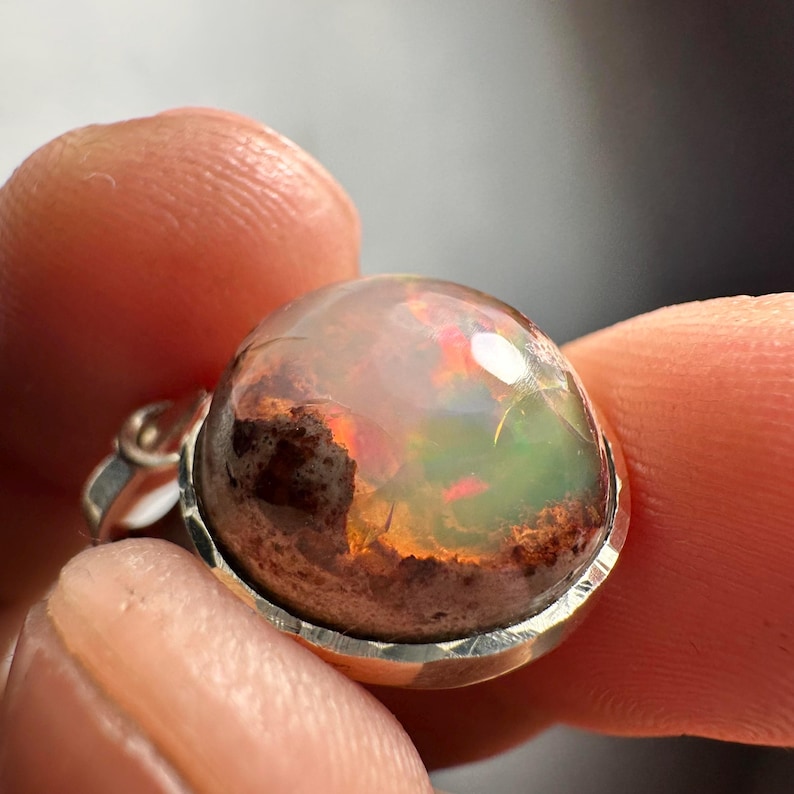 May include: A silver pendant with a round, iridescent opal stone. The opal has a brown, green, and orange color pattern.