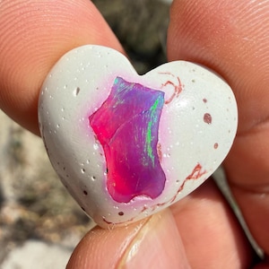 May include: A heart-shaped stone with a white exterior and a vibrant, multi-colored interior. The interior displays shades of pink, purple, and green. The stone has a textured surface with small holes and reddish-brown markings.