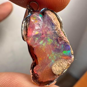 Op de afbeelding: Een zilveren hanger met een grote, ruwe opaal met een roze, oranje en groene iriserende glans. De opaal is gezet in een zilveren bezel en heeft een natuurlijke, ruwe uitstraling.