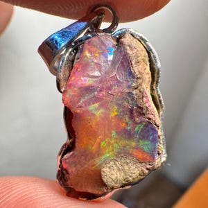 Op de afbeelding: Een ruwe opaal hanger met een zilveren setting. De opaal is een levendige roze, oranje en groene kleur met een ruwe, natuurlijke textuur.