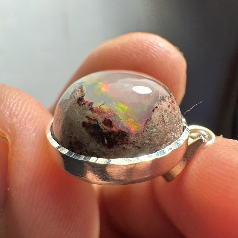 May include: A silver pendant with a round, iridescent opal stone set in a silver bezel. The opal has a white base with flashes of green, red, and orange.